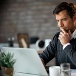 Man sitting at a desk in an office, looking at a laptop and thoughtfully touching his chin, suggesting concentration.
