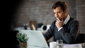 Man sitting at a desk in an office, looking at a laptop and thoughtfully touching his chin, suggesting concentration.