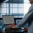 Person standing at a desk, typing on a laptop with a lighted keyboard in a dim control room or data center setting.