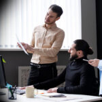 Three coworkers collaborate: a man in a beige shirt explains a document to a colleague in a wheelchair while a woman in a light blue blazer chats nearby.