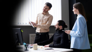Three coworkers collaborate: a man in a beige shirt explains a document to a colleague in a wheelchair while a woman in a light blue blazer chats nearby.