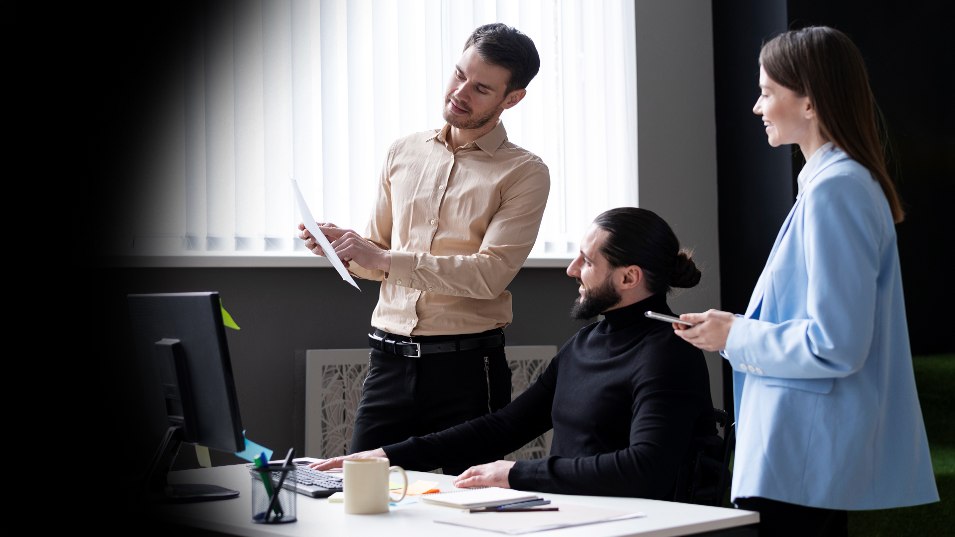 Three coworkers collaborate: a man in a beige shirt explains a document to a colleague in a wheelchair while a woman in a light blue blazer chats nearby.