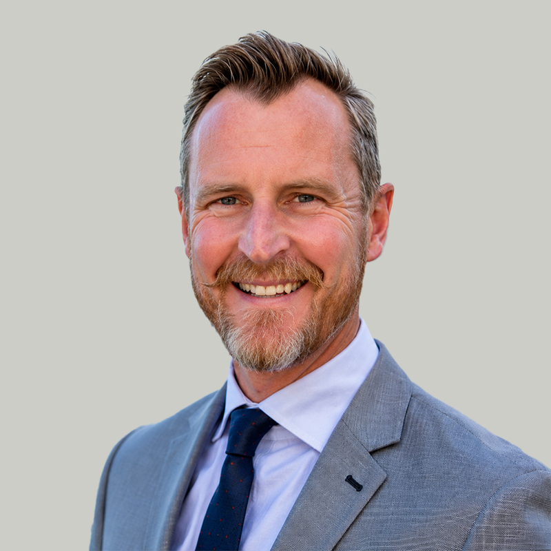 Smiling man in a light gray suit and navy tie, portrait against a neutral background.