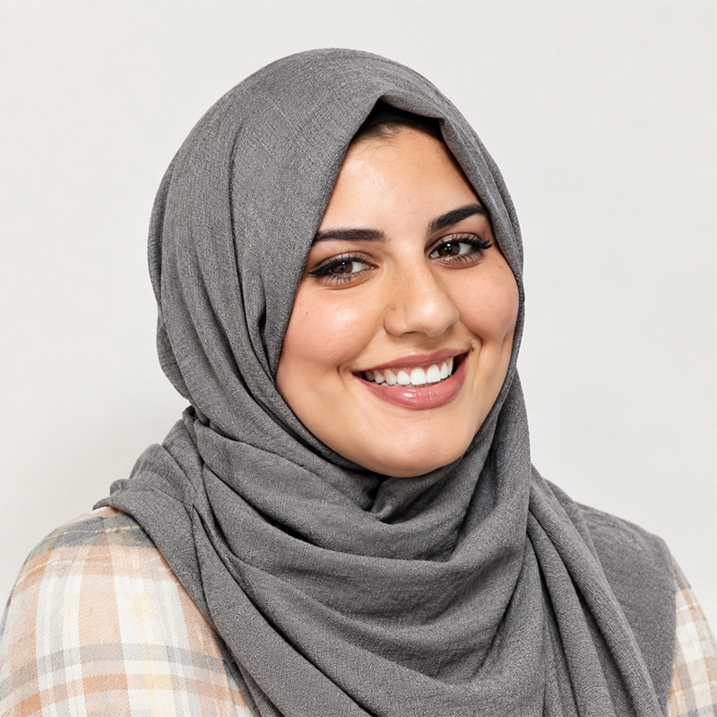 Smiling woman wearing a gray hijab and plaid shirt against a light background.